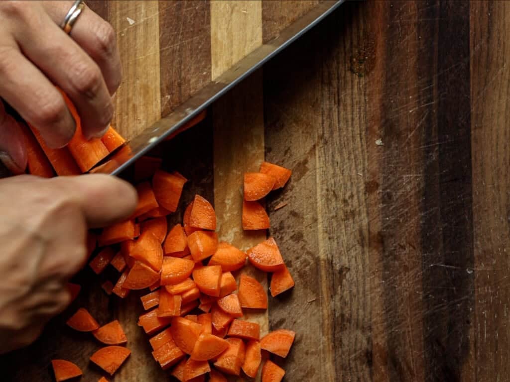 A close-up of hands slicing fresh carrots on a wooden cutting board with a large knife. The chopped carrot pieces are scattered on the board.